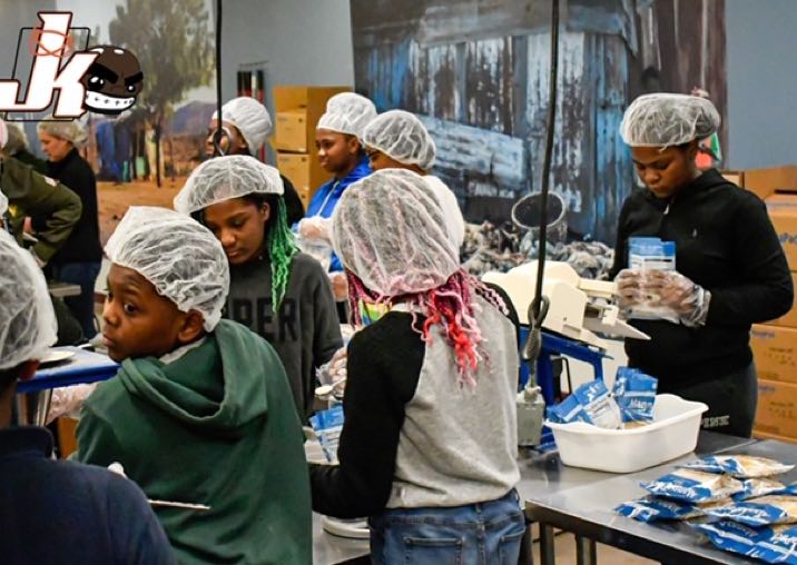 A room full of children wearing hair nets, packaging food packets. In the upper left of the photo, a logo reading "JK"