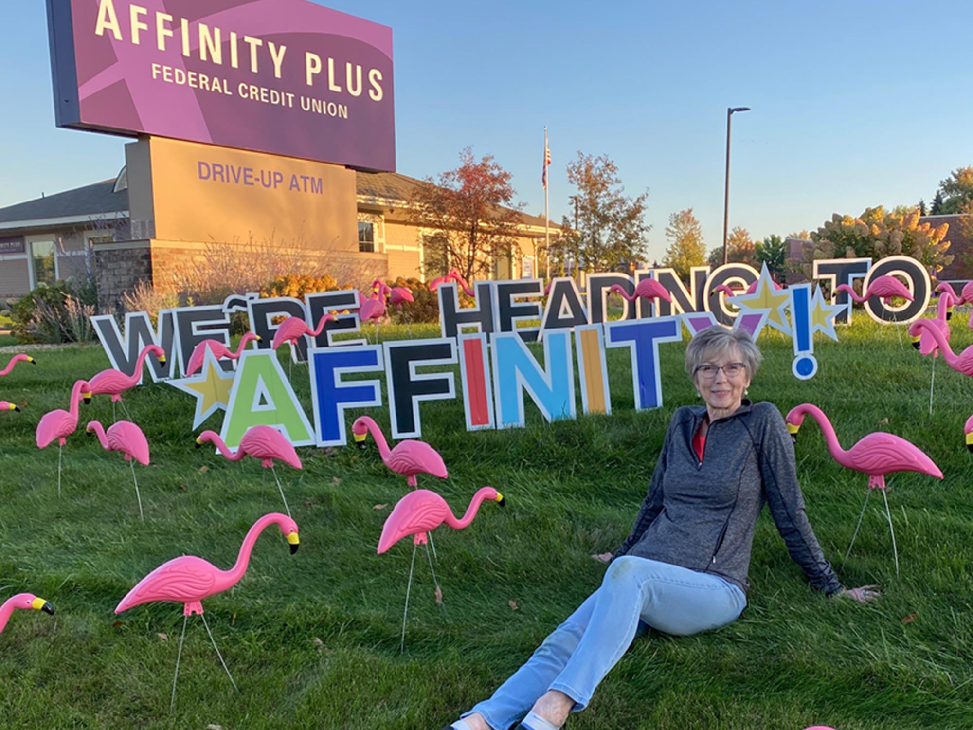 Woman sitting on the lawn in front of Affinity Plus branch with pink flamingos
