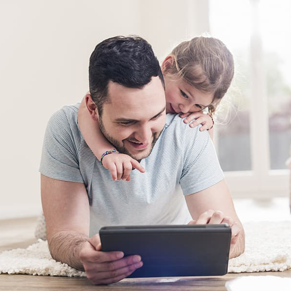A young girl hangs on her dad's shoulders to point and look at a tablet .